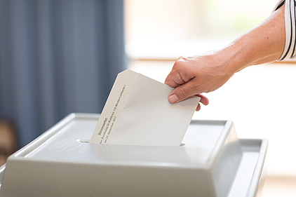 Eine Frauenhand steckt einen Briefumschlag in eine Wahlurne.